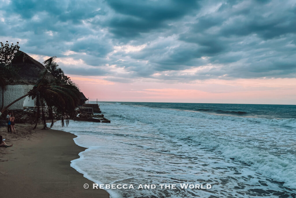Ocean waves roll onto a sandy beach beneath dramatic gray clouds tinged with pink light from the setting sun. A leaning palm tree and beachfront buildings frame the shoreline. Evenings like this capture the coastal charm of Palomino Colombia.