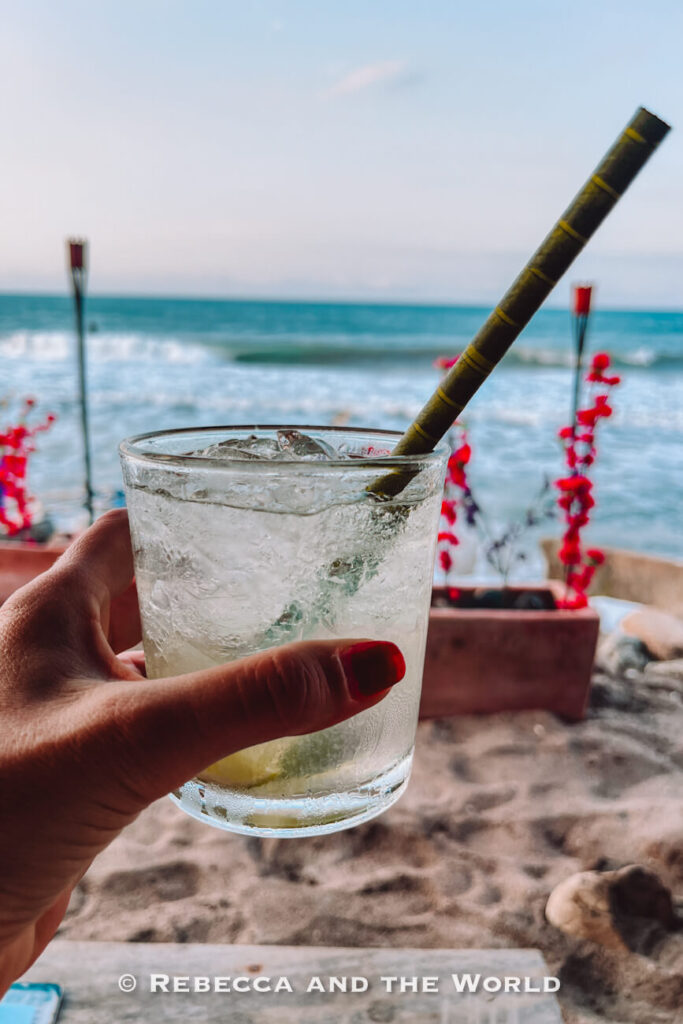 A hand holds a chilled cocktail filled with ice and a bamboo straw while the ocean stretches across the background. Bright red flowers and beachside seating add a tropical touch. Relaxing with a drink by the water is often recommended in a Palomino travel guide.