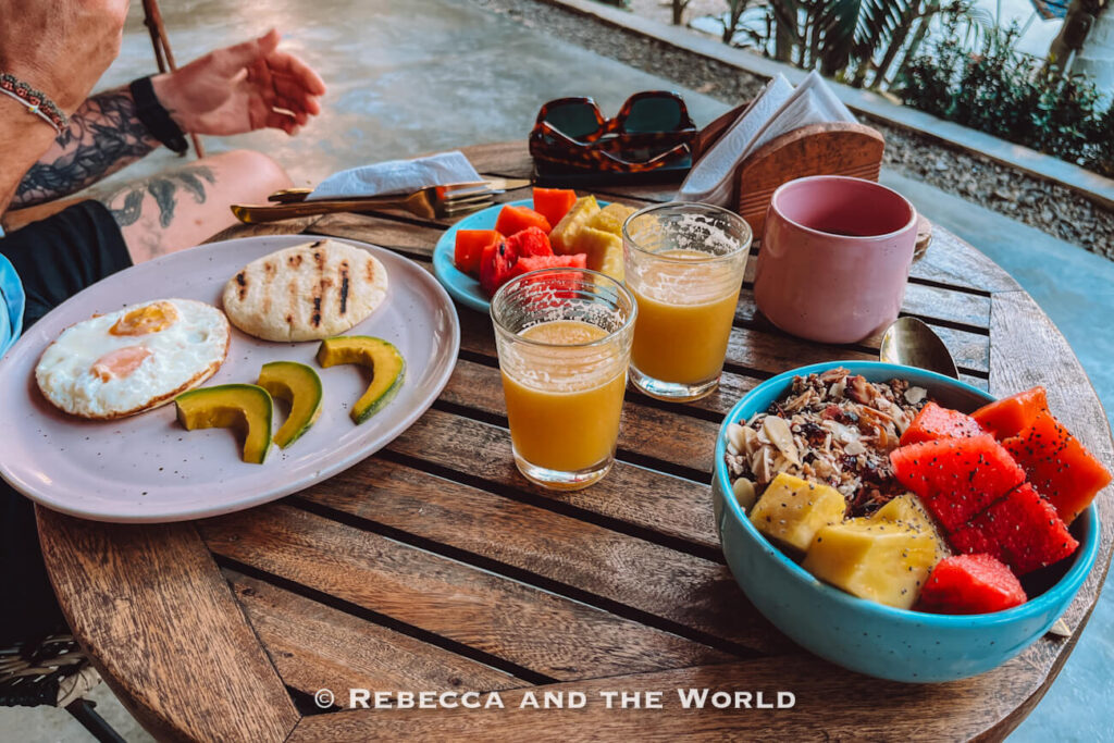 A wooden table is set with a colorful breakfast that includes fried eggs, sliced avocado, an arepa, fresh fruit, granola, and glasses of juice. A person sits nearby, enjoying a relaxed morning meal. Breakfasts like this are a highlight of many stays in Palomino Colombia.