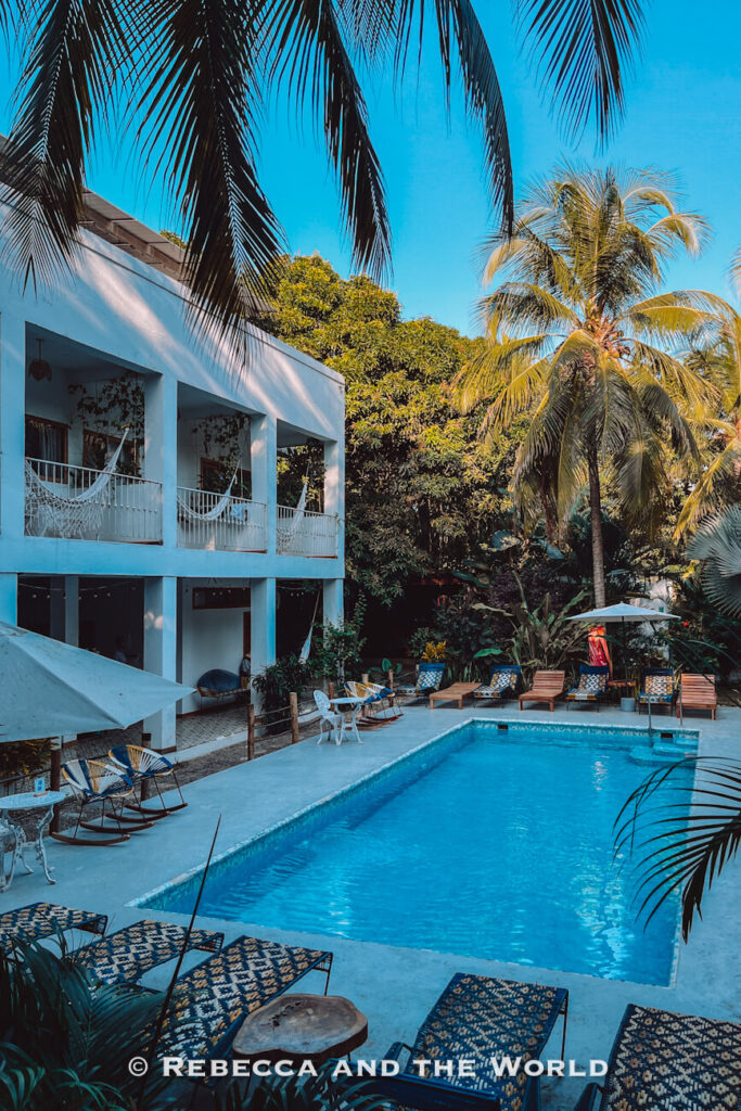 A long rectangular swimming pool sits in the center of a tropical courtyard framed by palm trees and lush greenery. White buildings with balconies and hammocks overlook the water, creating a tranquil atmosphere often featured in a Palomino travel guide.