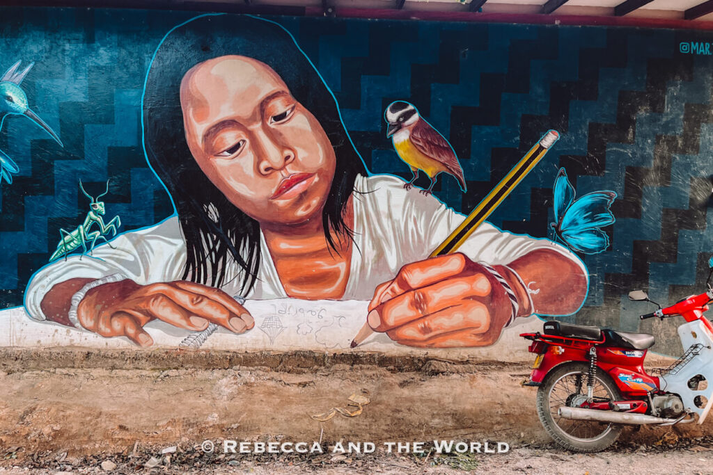 A large mural depicts an Indigenous girl drawing in a notebook, accompanied by a bird, butterfly, and praying mantis. A red motorcycle is parked nearby, emphasizing the scale of the artwork. Colorful murals like this add character to the streets of Palomino Colombia.