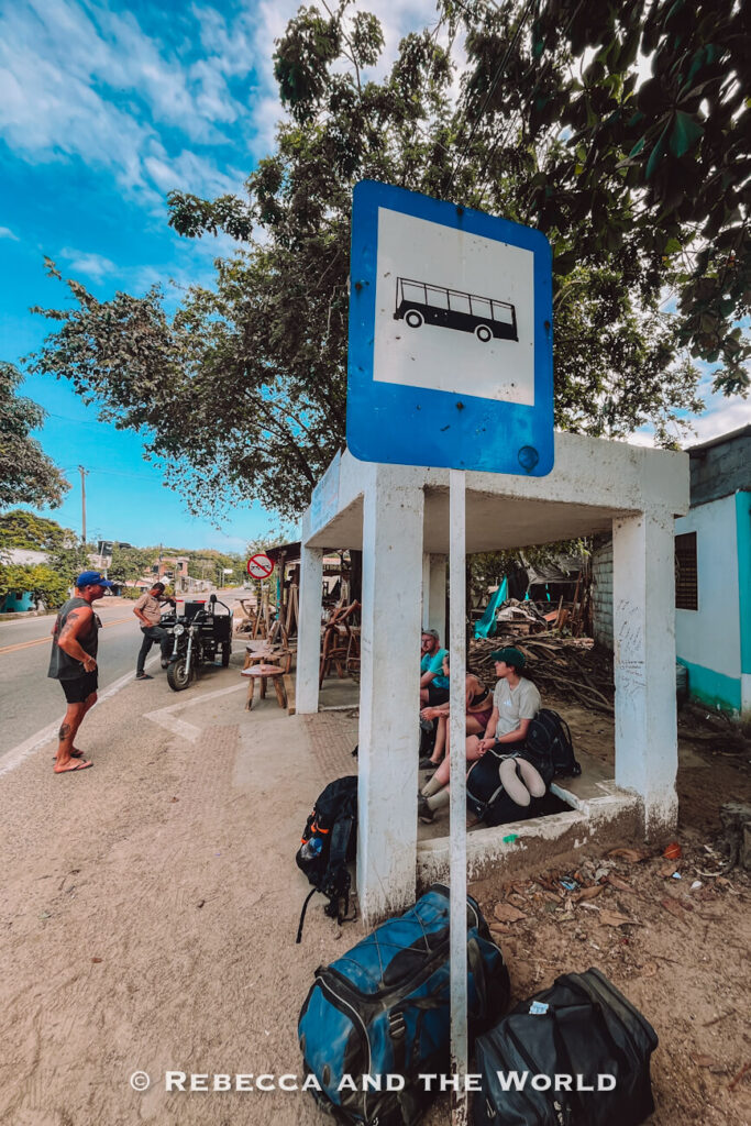 Travelers wait beneath a simple white concrete bus shelter with backpacks and luggage gathered around them. The roadside setting reflects the relaxed pace of transportation in the area. Stops like this are familiar sights for visitors following a Palomino travel guide.