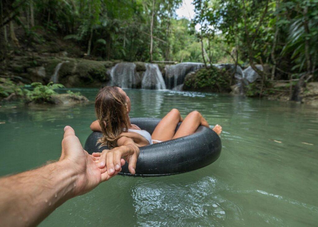 A person relaxes in an inner tube on calm green water while holding another person's hand. Small waterfalls cascade over rocks in the background, surrounded by dense tropical forest. Natural swimming spots like this are a popular answer to what to do in Palomino and nearby destinations.