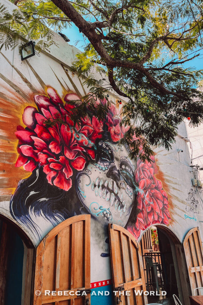 A large, colorful mural of a sugar-skull-style face framed by vibrant red flowers covers the white exterior wall of a colonial building. Wooden arched doors stand open beneath the artwork, while leafy tree branches stretch overhead against a bright blue sky. This striking urban artwork is one of the most photogenic things to do in Santa Marta.