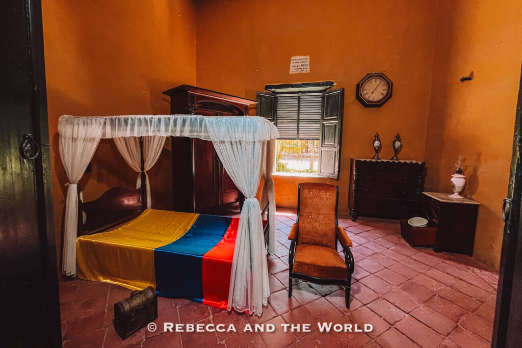 A preserved colonial-era bedroom with warm ochre walls features a wooden four-poster bed draped in white curtains. A Colombian flag-colored bedspread covers the mattress, while antique furniture, a rocking chair, and a wall clock complete the historic setting. This cultural attraction is where Simon Bolivar died and it offers insight into the history of Santa Marta Colombia.