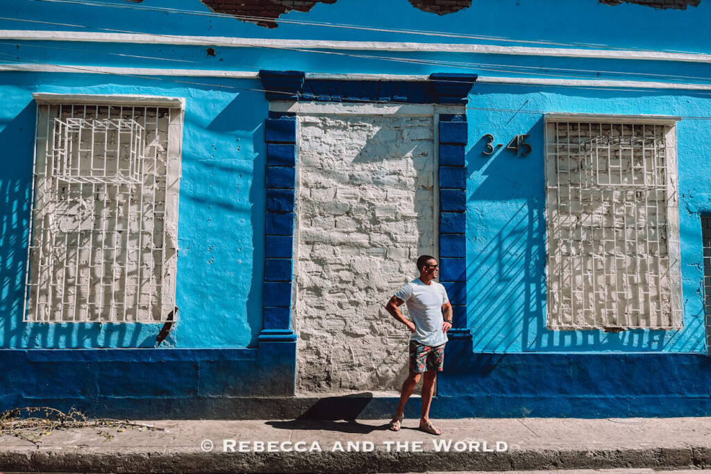 A vivid turquoise-blue colonial building with white iron window grilles stands along a quiet street. A casually dressed man - the author's husband - stands on the sidewalk in front of the building, creating contrast against the colorful architecture. Exploring streets like these is a highlight in many Santa Marta travel guide recommendations.