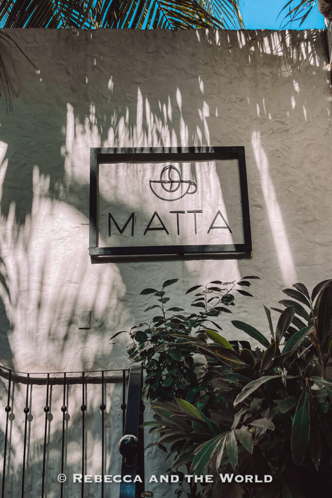 A minimalist sign reading “MATTA” hangs on a white stucco wall partially shaded by palm fronds. Tropical plants and a black metal gate frame the entrance, creating a peaceful courtyard atmosphere. Discovering hidden cafés is a popular answer to what to do in Santa Marta.