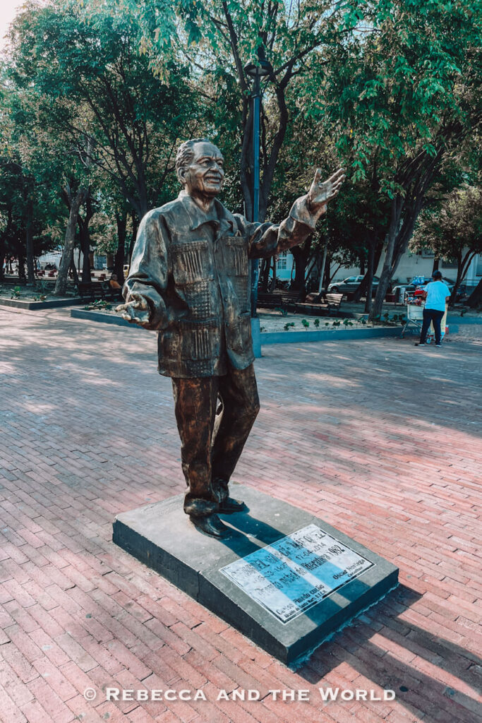 A bronze statue of a smiling man - Gabriel Garcia Marquez - stands on a stone pedestal in a tree-lined public square. The figure gestures with both hands while benches, pathways, and a street vendor appear in the background. Exploring local plazas is a worthwhile addition to any Santa Marta itinerary.