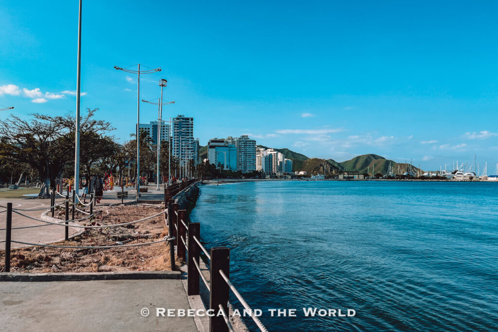 A paved seaside promenade runs alongside calm blue water with modern buildings, marina docks, and green hills visible in the distance. Tall light poles line the waterfront beneath a cloud-dotted sky. The marina district is frequently featured in every Santa Marta travel guide.