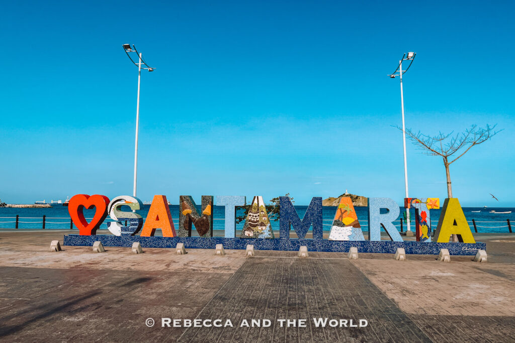 A large decorative “Santa Marta” sign sits along the waterfront promenade, with the Caribbean Sea and distant hills visible behind it. Each letter is decorated with colorful local artwork and patterns. Stopping for photos here is one of the classic answers to what to do in Santa Marta.