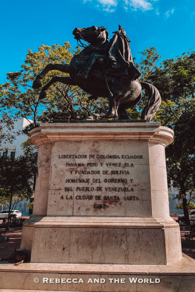 A dramatic bronze equestrian statue stands atop a large stone pedestal engraved with historical text. The rearing horse and rider are silhouetted against a clear blue sky and surrounded by trees. Historical monuments like this - a statue of Simon Bolivar - are among the most interesting things to do in Santa Marta.