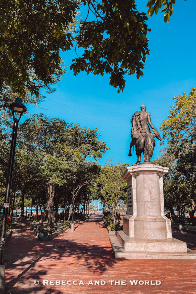 A brick pathway leads through a leafy urban park toward the waterfront. An equestrian monument rises on the right side, while mature trees provide shade throughout the landscaped square. This scenic area - Parque Bolivar - is a peaceful stop during a Santa Marta Colombia sightseeing day.