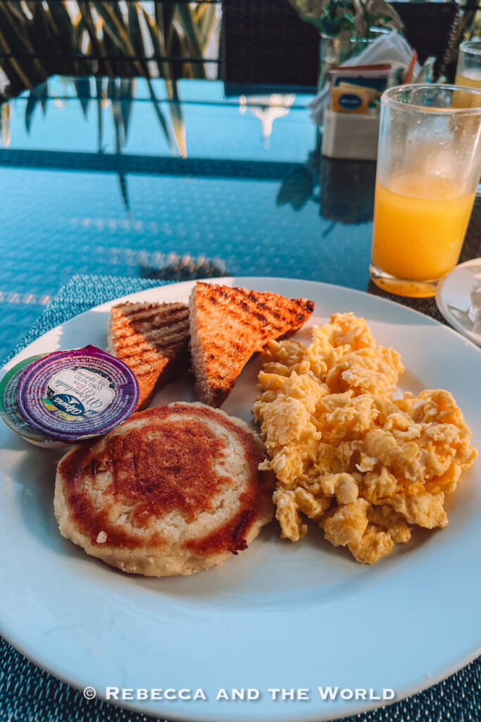 A white plate holds scrambled eggs, toasted bread triangles, and a golden arepa, accompanied by a glass of orange juice. The meal sits on a glass-topped table in a bright dining area. Sampling local breakfasts is often recommended in a Santa Marta travel guide.