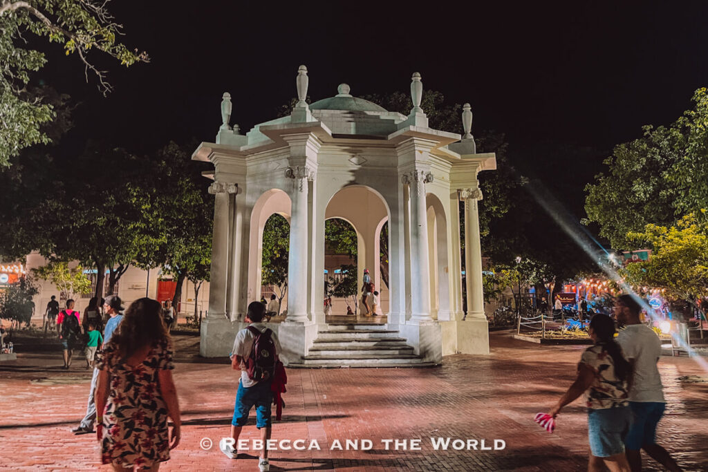 A white neoclassical gazebo with columns and arched openings stands in the center of a brick-paved plaza at night. People stroll around the structure while warm lights illuminate the square and surrounding trees against a dark sky. Evening walks through plazas like this are among the most enjoyable things to do in Santa Marta.