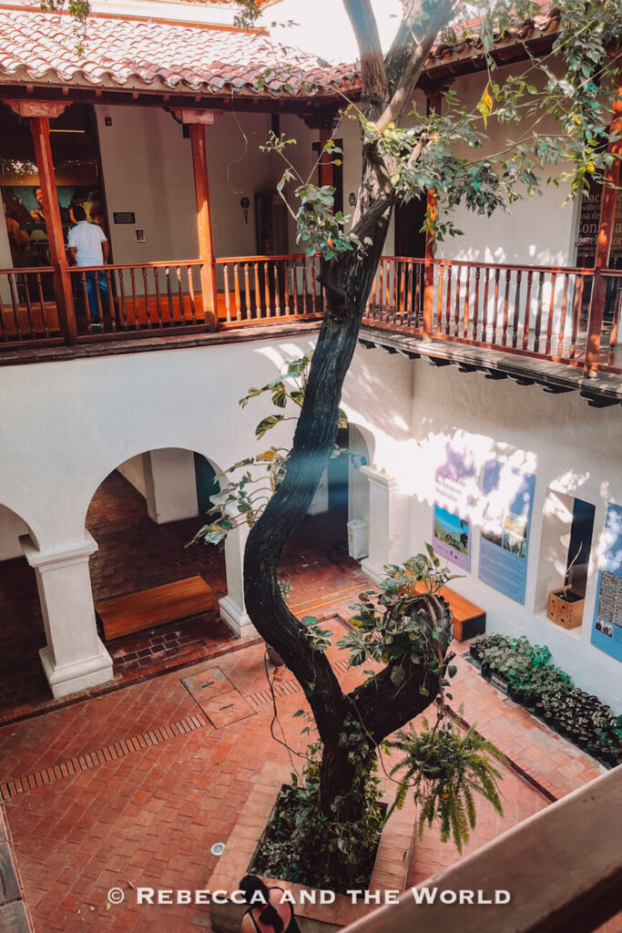 A two-story colonial courtyard features white walls, wooden balconies, brick floors, and a large tree growing from the center. Sunlight filters through the open roof, casting soft shadows across the peaceful interior space. Museo del Oro Tairona is a must-visit on a Santa Marta itinerary.