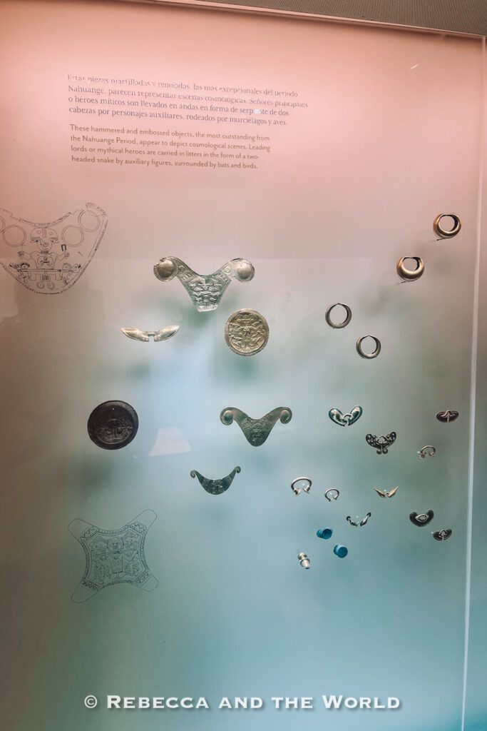 A glass museum exhibit showcases small gold and metal ornaments, rings, and decorative objects arranged against a pale background. Informational text and illustrations explain their cultural significance and archaeological context. Visiting museums is a rewarding way to learn more about Santa Marta Colombia.