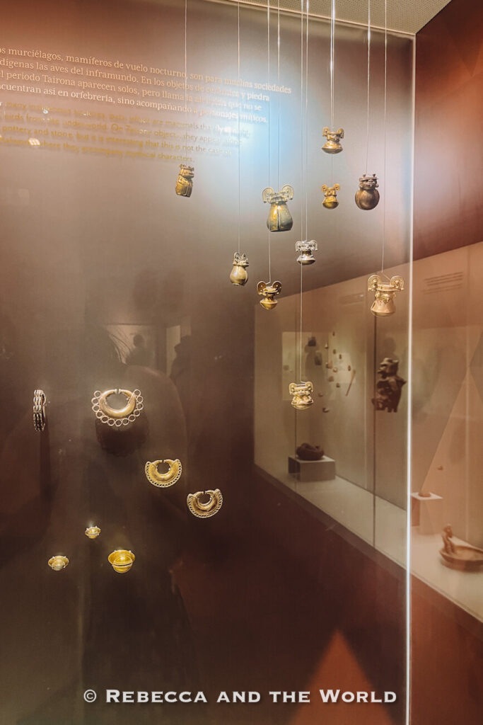 Delicate gold ornaments and ceremonial objects hang inside a museum display case, illuminated against a dark backdrop. Additional exhibits and interpretive panels can be seen reflected in the glass. Exploring Indigenous history exhibits is one of the more educational things to do in Santa Marta.