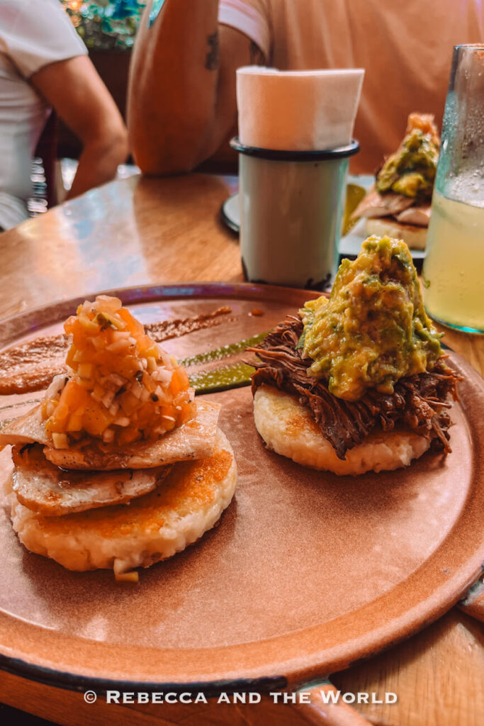 Two small round corn arepas are topped with savory fillings, including shredded meat, guacamole, grilled fish, and fresh tomato salsa. The dish is served on a ceramic plate at a wooden restaurant table beside a cold drink. Sampling local cuisine is often featured in every Santa Marta travel guide.