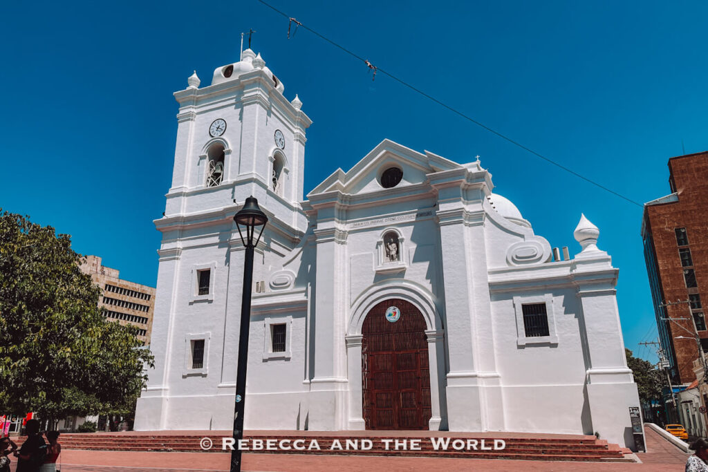 A large white cathedral with a clock tower, arched entrance, and classical architectural details stands in an open plaza beneath a vivid blue sky. Wide brick steps lead to the wooden front doors. Visiting landmarks like this is an essential part of any Santa Marta itinerary.