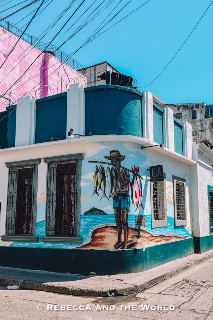 A colorful mural painted on the corner of a building depicts a fisherman carrying freshly caught fish beside the sea. The artwork wraps around the structure and reflects the city’s coastal identity. Street art celebrating local culture is one of the highlights of Santa Marta Colombia.
