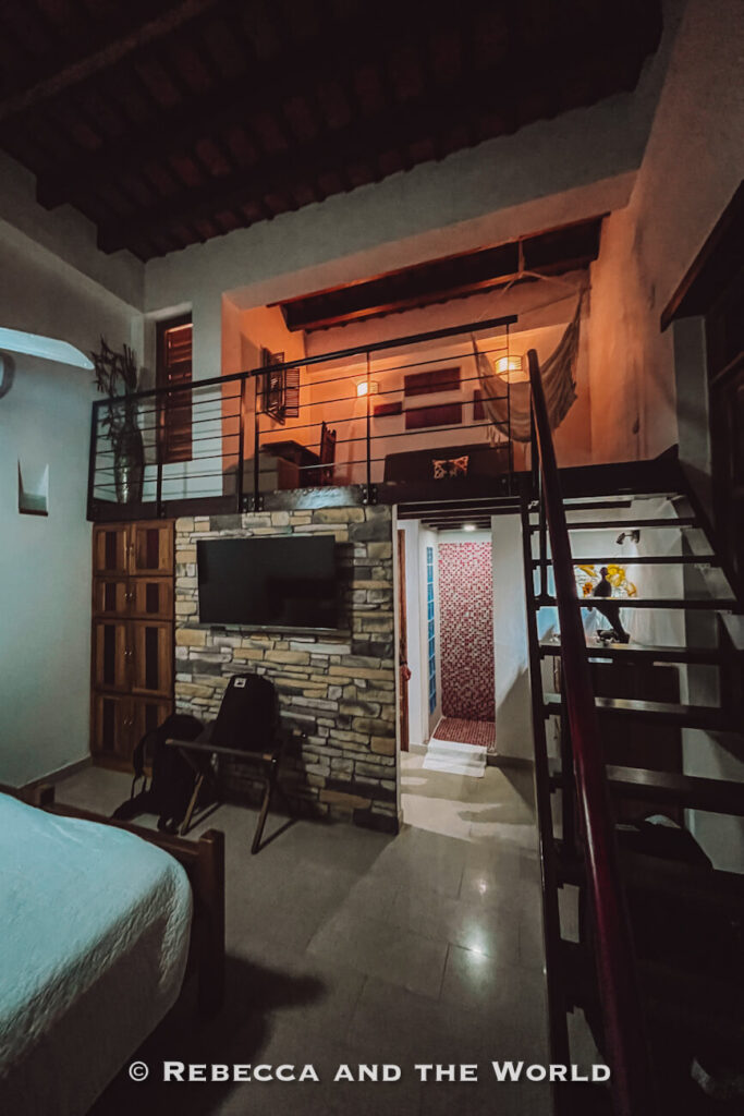 A cozy loft-style room features a bed, stone accent wall, television, staircase, and elevated sleeping area with warm lighting. Exposed wooden ceiling beams and rustic décor create a welcoming atmosphere. Boutique accommodations like this can be a comfortable addition to a Santa Marta itinerary.
