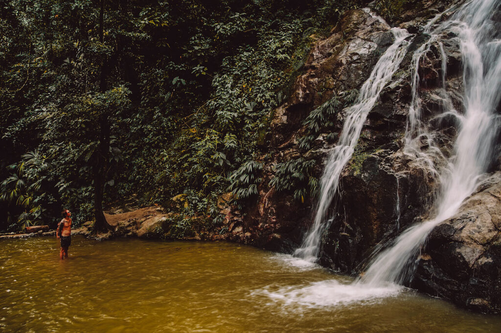 A cascading waterfall flows down dark rock formations into a shallow natural pool surrounded by dense tropical rainforest. A person stands in the water near the edge of the falls, emphasizing the scale of the lush jungle setting. Visiting hidden waterfalls like this one in Minca is one of the most adventurous things to do in Santa Marta.