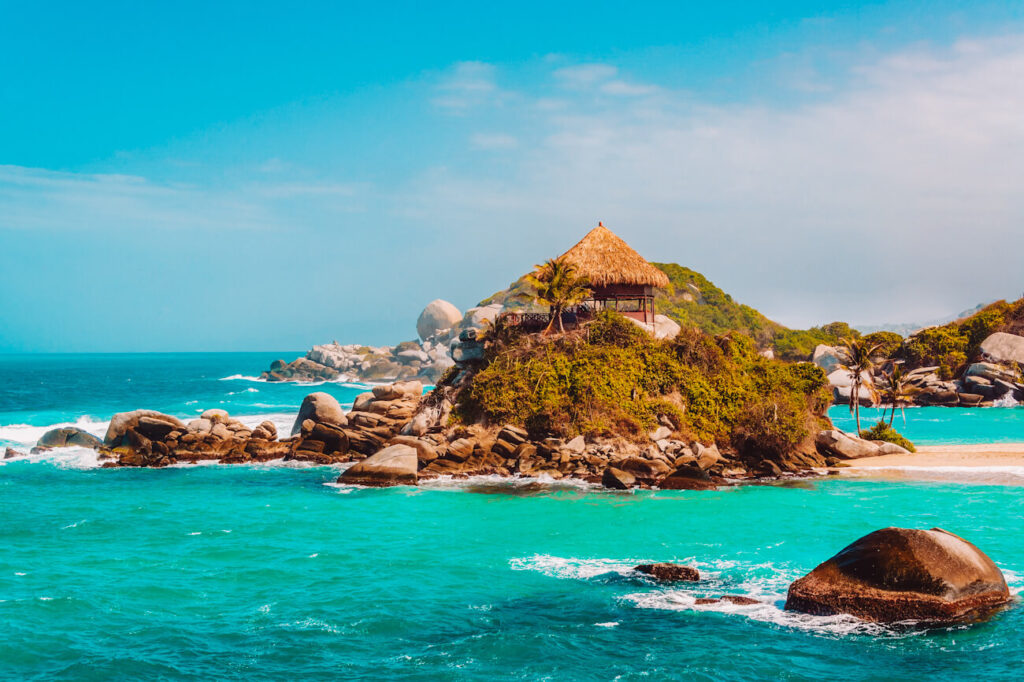 A small thatched-roof hut sits atop a rocky outcrop covered in greenery, surrounded by turquoise Caribbean waters and giant granite boulders. Waves crash against the shoreline while a bright blue sky stretches across the horizon, creating a postcard-perfect coastal scene. Taking a day trip from Santa Marta to Tayrona National Park is a great way to see this beautiful setting.