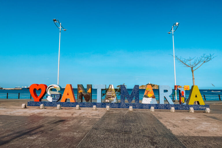 A large decorative “Santa Marta” sign sits along the waterfront promenade, with the Caribbean Sea and distant hills visible behind it. Each letter is decorated with colorful local artwork and patterns. Stopping for photos here is one of the classic answers to what to do in Santa Marta.