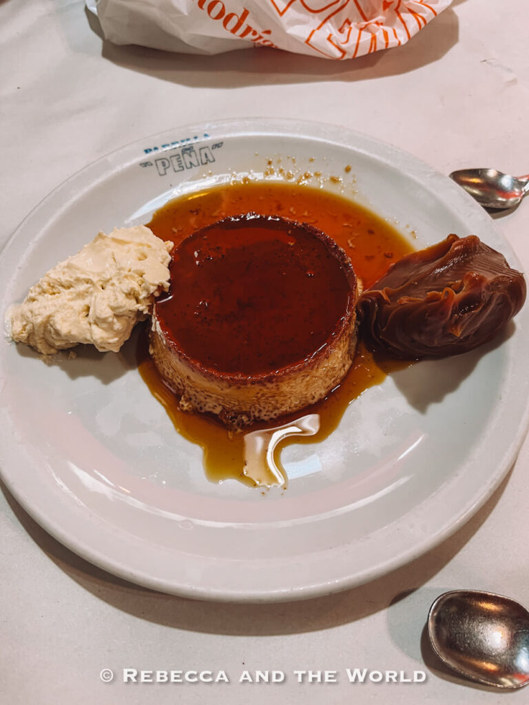 A plate of caramel flan topped with glossy syrup sits on a white table, served with a scoop of cream and a dollop of dulce de leche at Parrilla Peña, one of the best steakhouses in Buenos Aires. Two metal spoons rest beside the dessert plate, and a plastic takeaway bag is partially visible in the background.