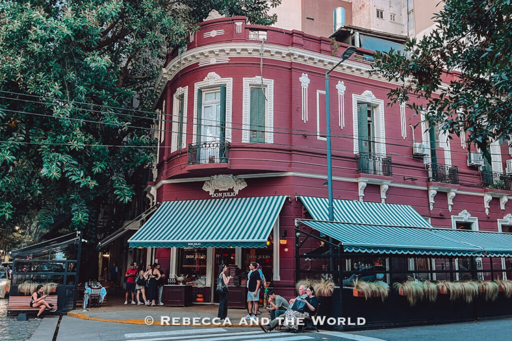 Exterior view of Don Julio, one of the best steakhouses in Buenos Aires, with its deep red corner building, striped awnings, and people gathered outside beneath leafy trees in Palermo.