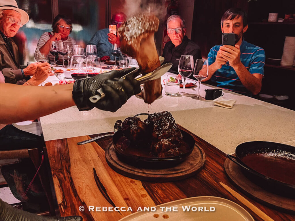 Guests seated around an open kitchen counter at The Argentine Experience while a chef serves smoked grilled meat, with several diners photographing the presentation during one of the popular things to do in Buenos Aires. The chef is pulling the bone cleanly out of a dish of osso bucco.