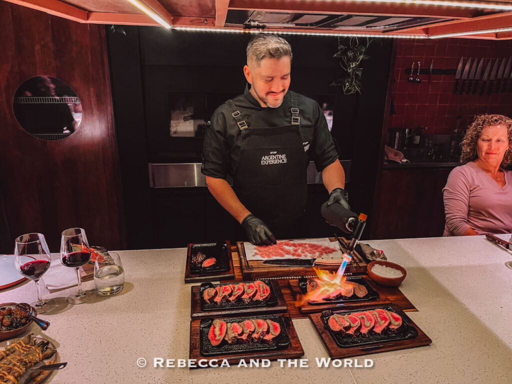 Chef at The Argentine Experience using a blowtorch to finish sliced steak dishes in an intimate open-kitchen dining space in Buenos Aires.