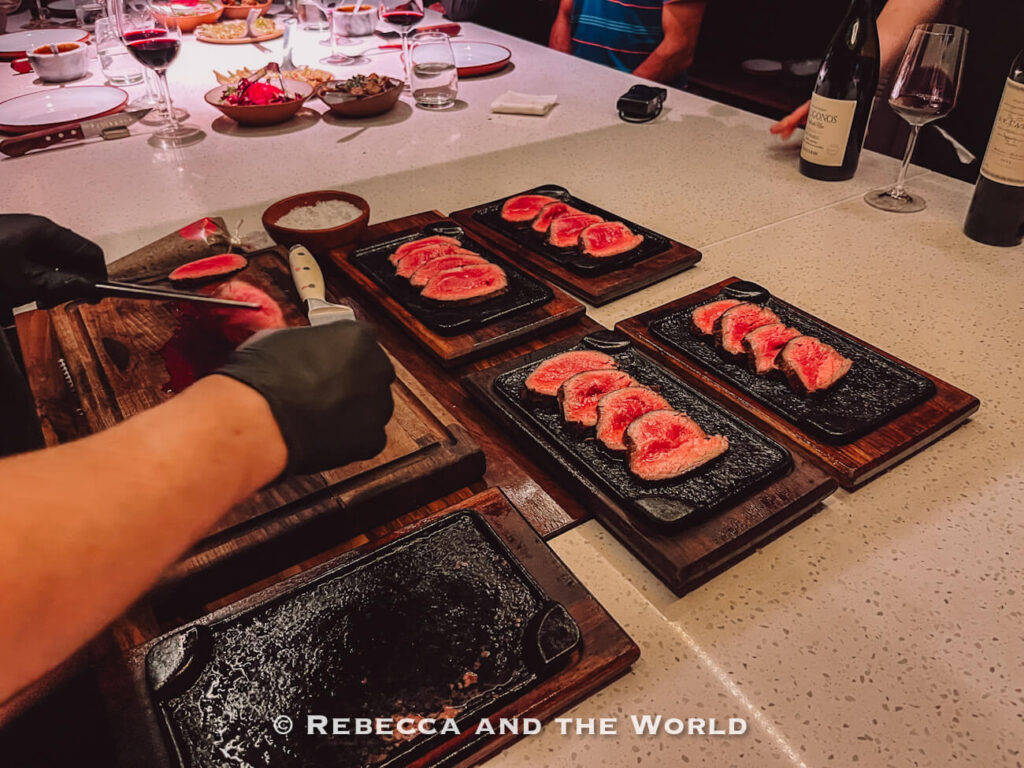 Close-up of sliced medium-rare steak plated on hot serving stones at The Argentine Experience, with wine glasses and shared dishes visible around the dining counter.