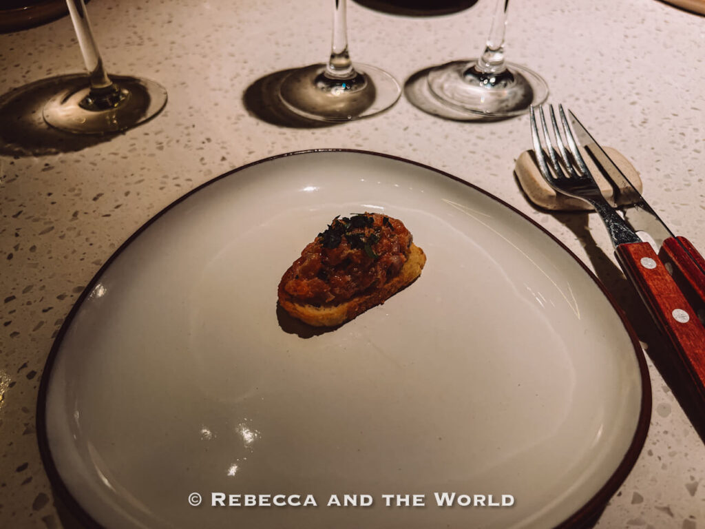 Bite-sized appetizer served on toasted bread in the center of a large white plate, with wine glasses and steak knives arranged on the dining counter at The Argentine Experience.