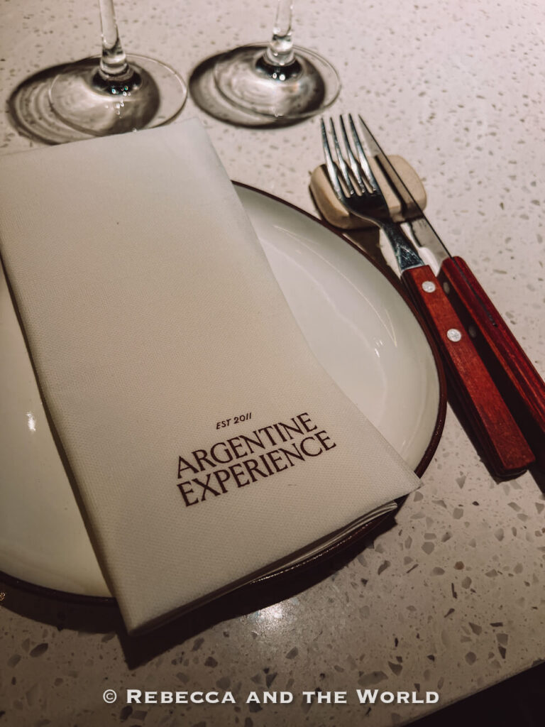 Table setting at The Argentine Experience featuring branded napkin, steak knife, fork, wine glasses, and ceramic dinner plate on a white stone counter.