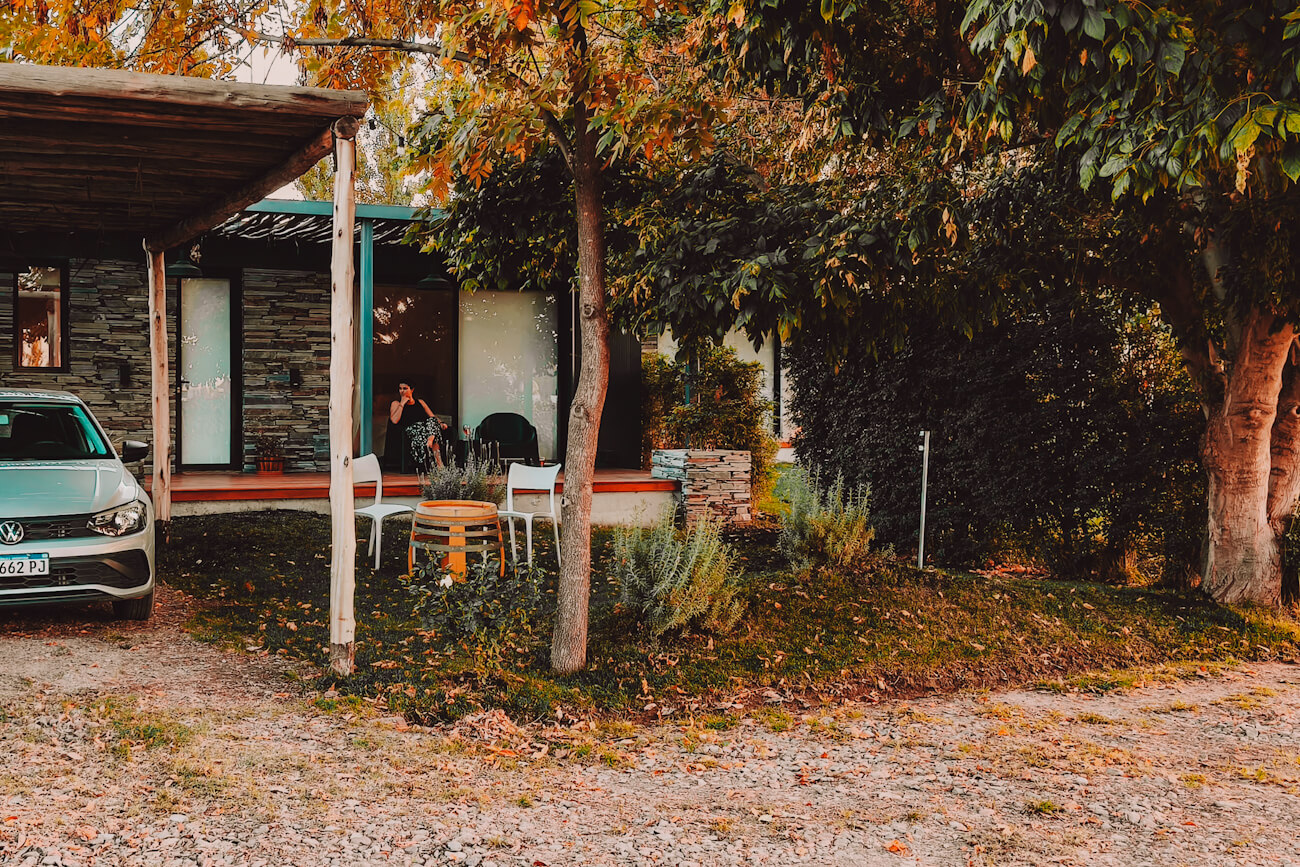 Small modern cabin surrounded by mature trees and autumn leaves, with a gravel driveway, parked white car, shaded patio seating, and a person sitting in the doorway relaxing outdoors. A tranquil accommodation setting and inspiration for what to do in Mendoza.