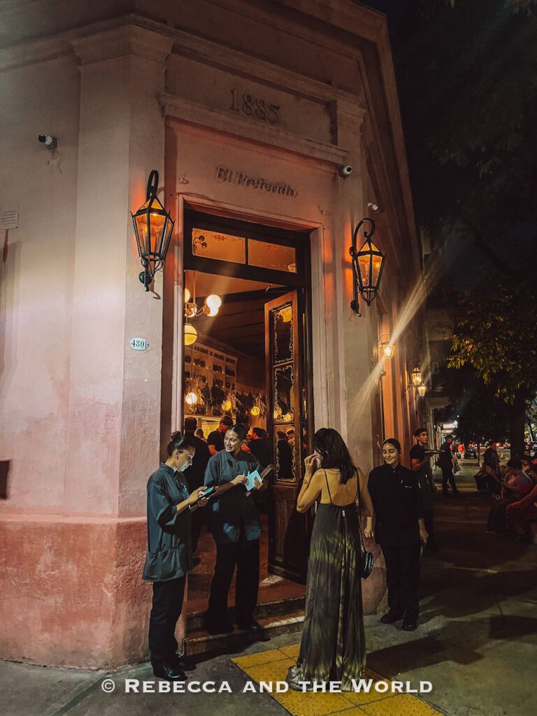 Nighttime street view of the entrance to a historic restaurant in Buenos Aires (El Preferido de Palermo), with tall lantern-style lights mounted on a pale pink building marked “1885.” Several staff members stand outside greeting guests while diners gather near the open doorway. Warm interior lighting spills onto the sidewalk, creating a lively evening atmosphere at one of the best places to eat in Buenos Aires.