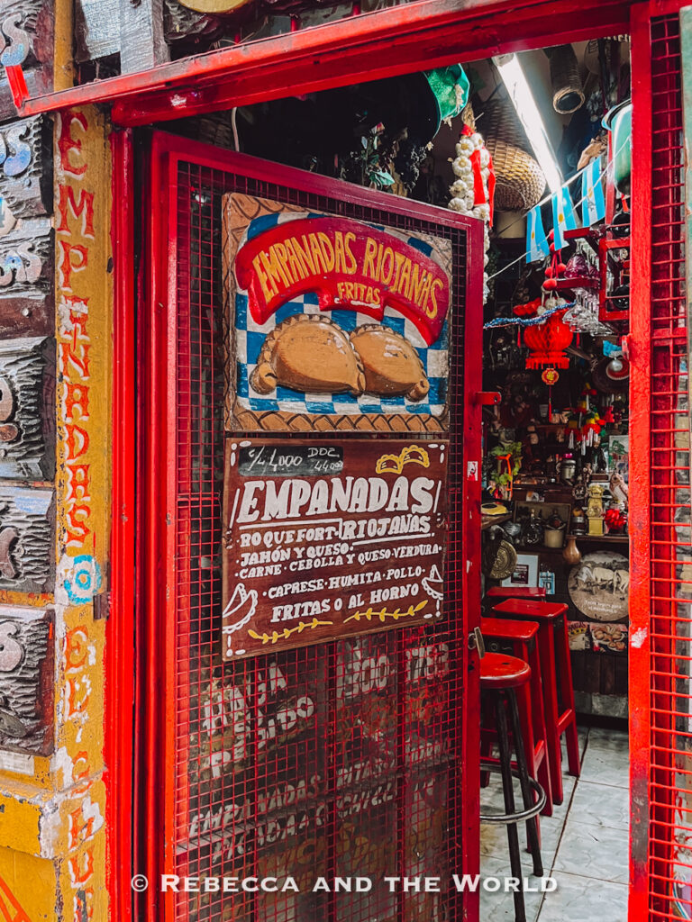 Narrow red-painted storefront selling traditional empanadas in Buenos Aires, with hand-painted signs advertising “Empanadas Riojanas.” The interior of El Gauchito is packed with decorations, stools, hanging lanterns, and Argentine flags, capturing the colorful local food scene and authentic Buenos Aires restaurants.