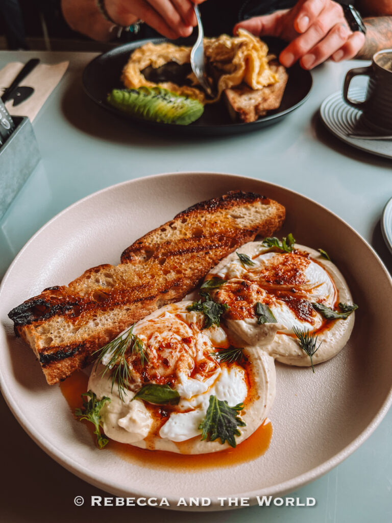 Close-up of a plated brunch dish with creamy poached eggs topped with herbs and chili oil, served with thick slices of toasted bread. Another breakfast plate and coffee sit blurred in the background, showcasing stylish café dining and where to eat in Buenos Aires for brunch.