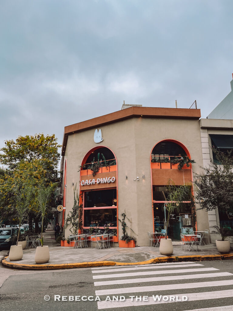 Exterior of Casa Dingo café on a street corner in Buenos Aires, featuring large arched windows, beige walls, orange trim, outdoor seating, and cloudy skies overhead. The modern café design highlights trendy Buenos Aires restaurants and cafés.