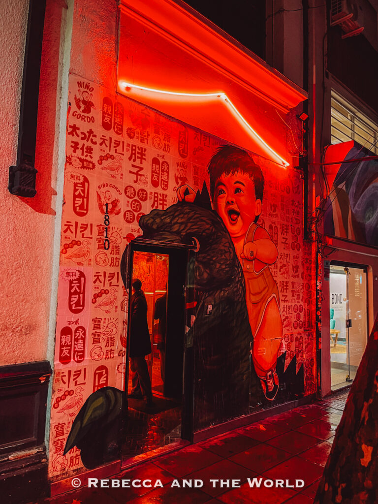 Bright red neon-lit restaurant entrance covered in illustrated posters and Asian-inspired graphics. A large mural of a smiling child surrounds the doorway while the wet sidewalk reflects the glowing lights, creating a dramatic nighttime dining scene in Buenos Aires.