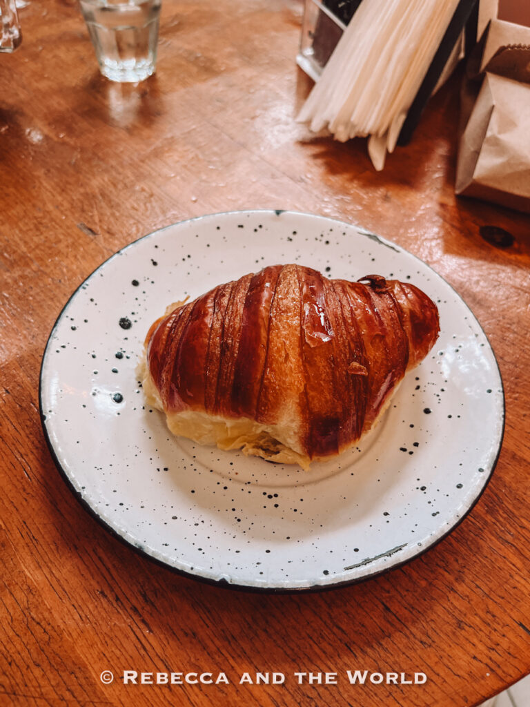 Golden-brown croissant served on a white speckled plate atop a rustic wooden table inside a café. The flaky pastry is photographed close-up, representing popular bakery stops and the best places to eat in Buenos Aires for pastries and coffee.