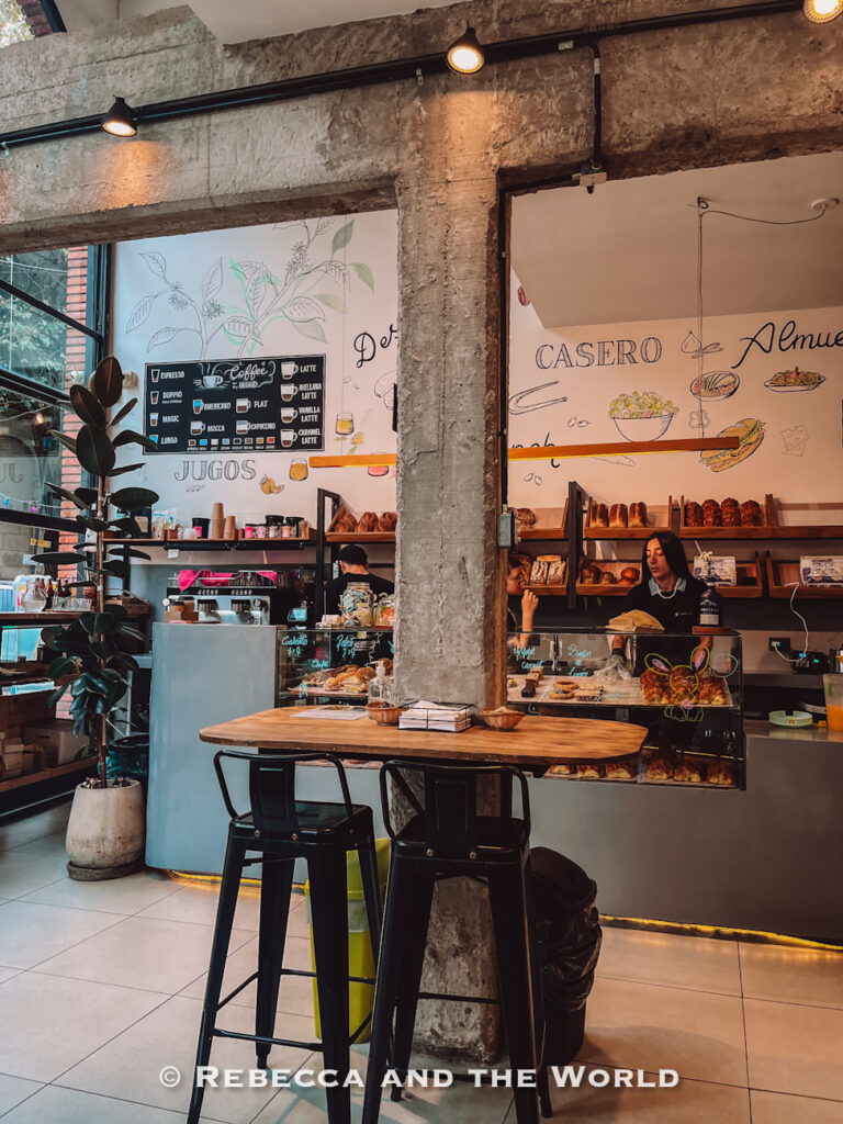 Interior of a modern bakery café with concrete pillars, hanging lights, shelves of bread and pastries, and a service counter staffed by employees. Hand-drawn menu art decorates the walls, capturing the relaxed café culture and where to eat in Buenos Aires for breakfast.