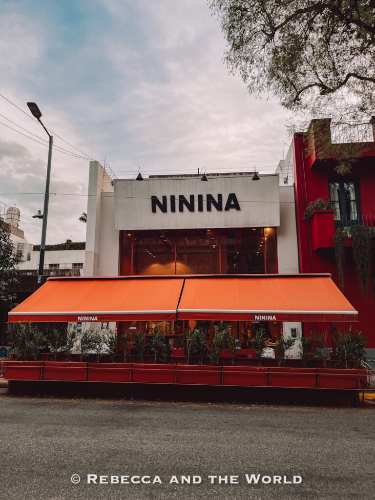 Exterior of the restaurant Ninina in Buenos Aires, with large black lettering above bright orange awnings and a red outdoor seating area. The modern storefront stands along a tree-lined street, highlighting one of the stylish places where to eat in Buenos Aires.