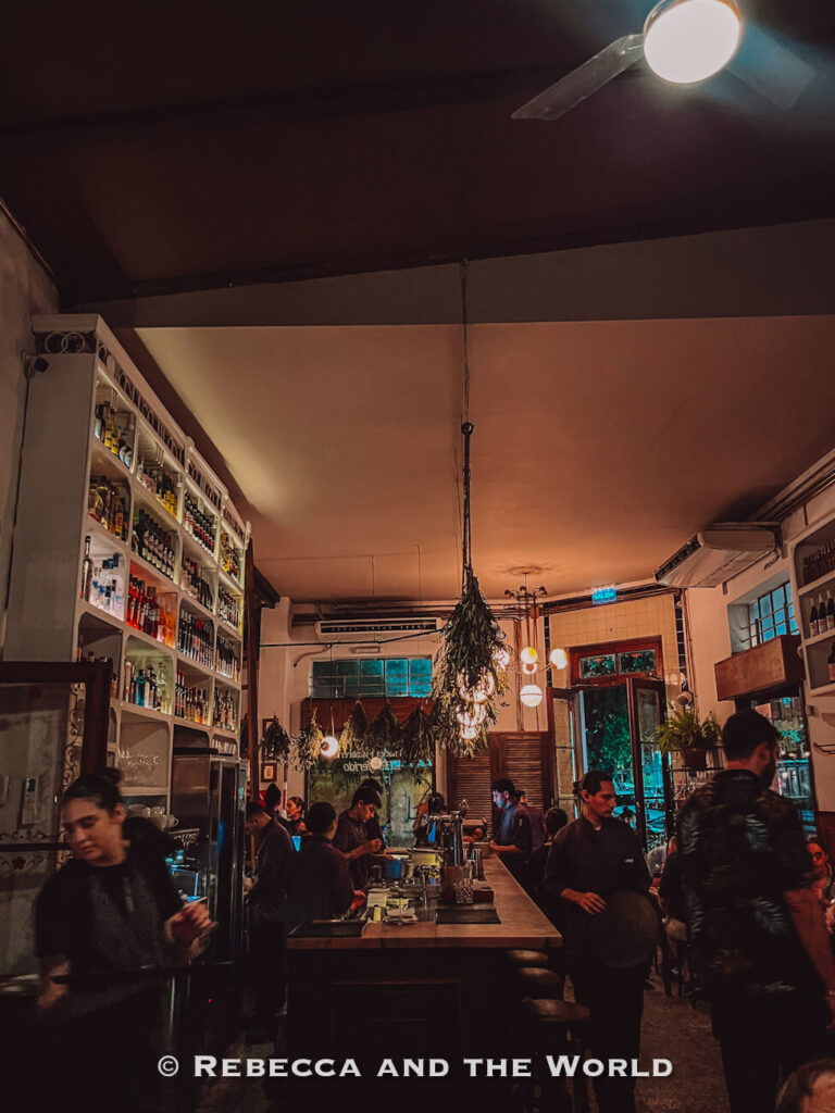Interior of a busy restaurant with high ceilings, hanging greenery, white shelving filled with bottles, and staff working behind a long marble bar. Diners sit at tables throughout the warmly lit dining room, illustrating the lively atmosphere of Buenos Aires restaurants.