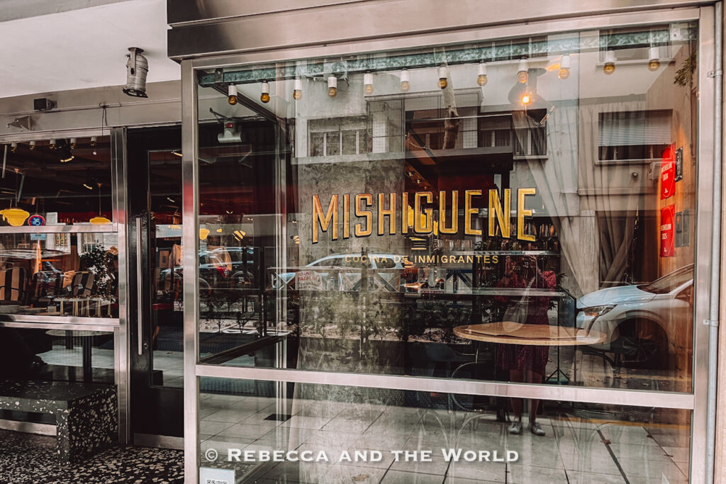 Glass-fronted entrance to Mishiguene restaurant in Buenos Aires, with gold lettering reading “Mishiguene Cocina de Inmigrantes.” Reflections of the street and nearby cars appear in the windows of the upscale dining space.