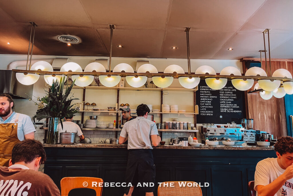 Bright café interior with globe pendant lights hanging above an open kitchen and espresso bar. Staff prepare food behind the counter while diners sit at wooden tables, showcasing one of the best places to eat in Buenos Aires for brunch or coffee.