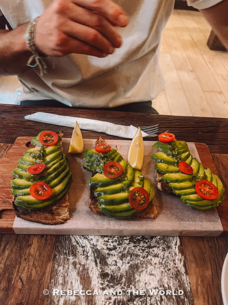 Wooden serving board topped with three slices of avocado toast layered with sliced avocado, cherry tomatoes, and green sauce. Lemon wedges sit between the toasts while a diner reaches across the table, highlighting popular brunch food in Buenos Aires restaurants.