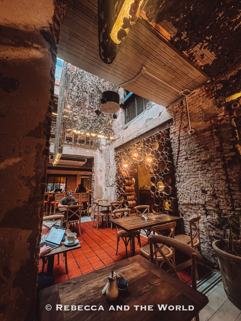 Rustic café courtyard with exposed brick walls, warm lighting, wooden tables, and decorative circular shelving made from stacked pipes. Guests work on laptops and relax with coffee in the cozy indoor-outdoor seating area, representing charming hidden spots where to eat in Buenos Aires.