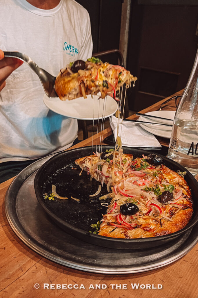 Close-up of a cheesy pizza slice being lifted from a black pan, with melted cheese stretching from the pie. The pizza is topped with olives, onions, and herbs, photographed inside a dimly lit restaurant known for comforting food and some of the best places to eat in Buenos Aires.